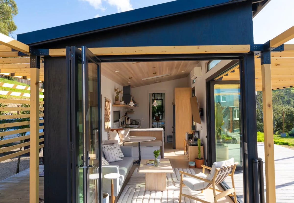 Modern Tiny House with Open Glass Doors, Cozy Interior Featuring Wooden Accents, Minimalist Furniture, and Bright Natural Light Streaming into Living and Kitchen Area Surrounded by Lush Greenery